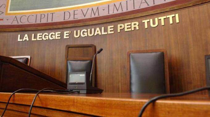Italian courtroom bench with microphones and empty chairs, wall text reads 'LA LEGGE E UGUALE PER TUTTI'.