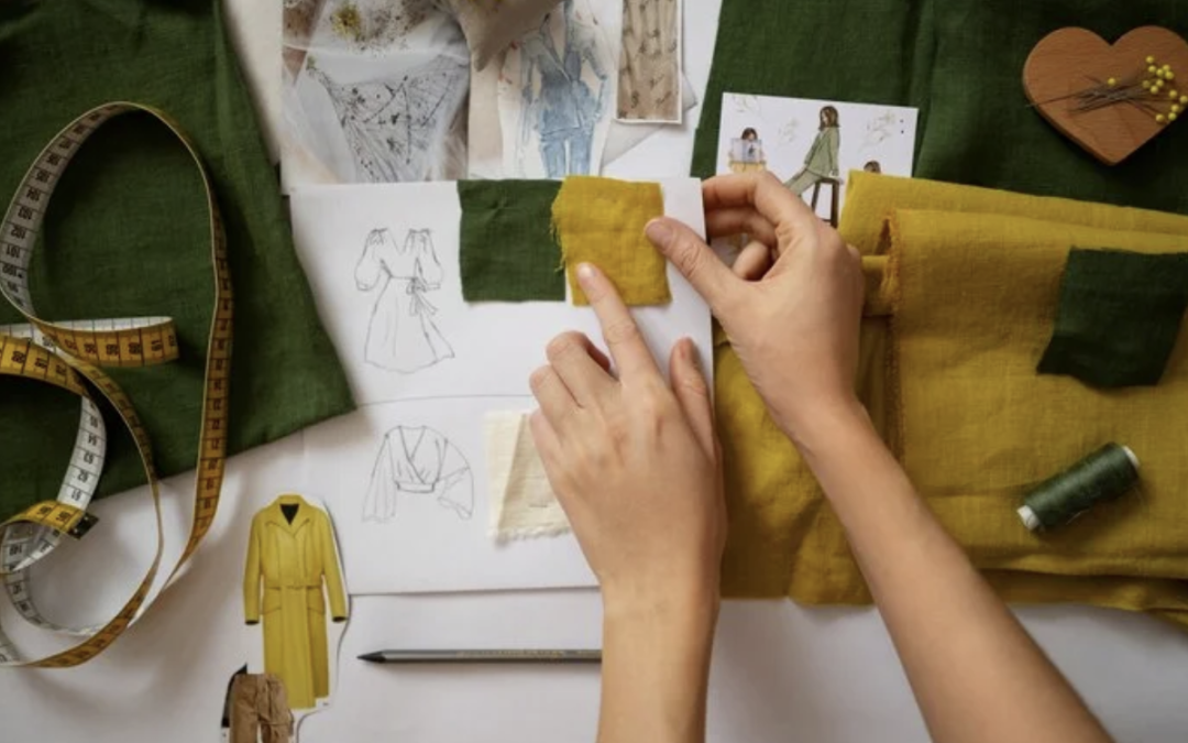 Fashion designer at a workspace, placing mustard fabric swatch on a sketchbook with clothing drawings and fabric nearby.